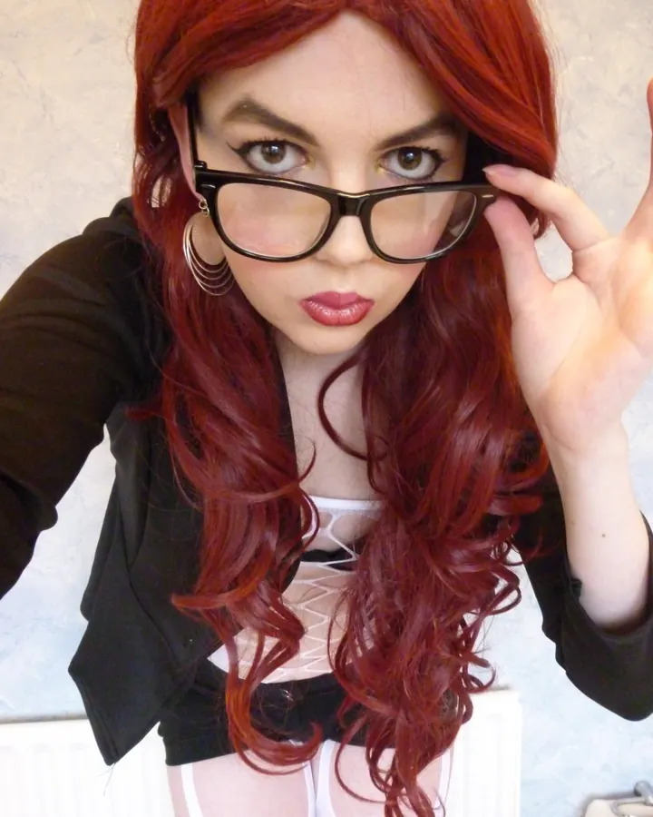 Femme aux cheveux roux, lunettes noires, vêtue d’un blazer noir et d’un soutien-gorge blanc, posant en intérieur.