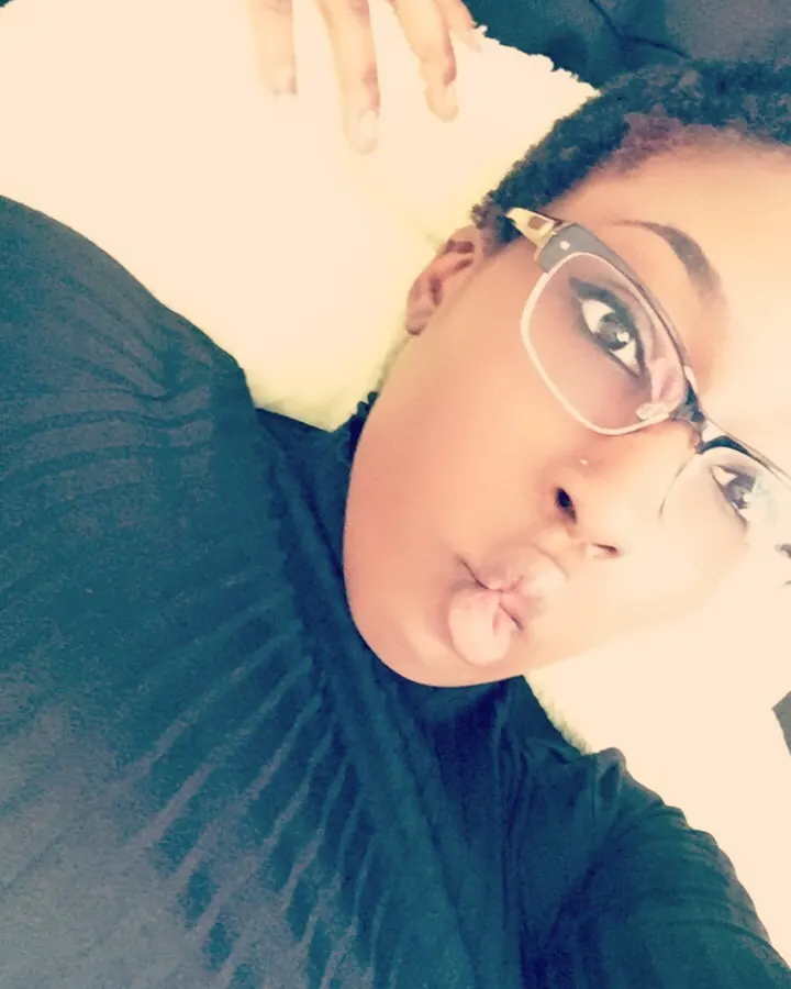 Femme africaine aux cheveux courts, vêtue d'un haut noir, assise sur un lit, dos tourné, regard vers l'appareil, cheveux mouillés.