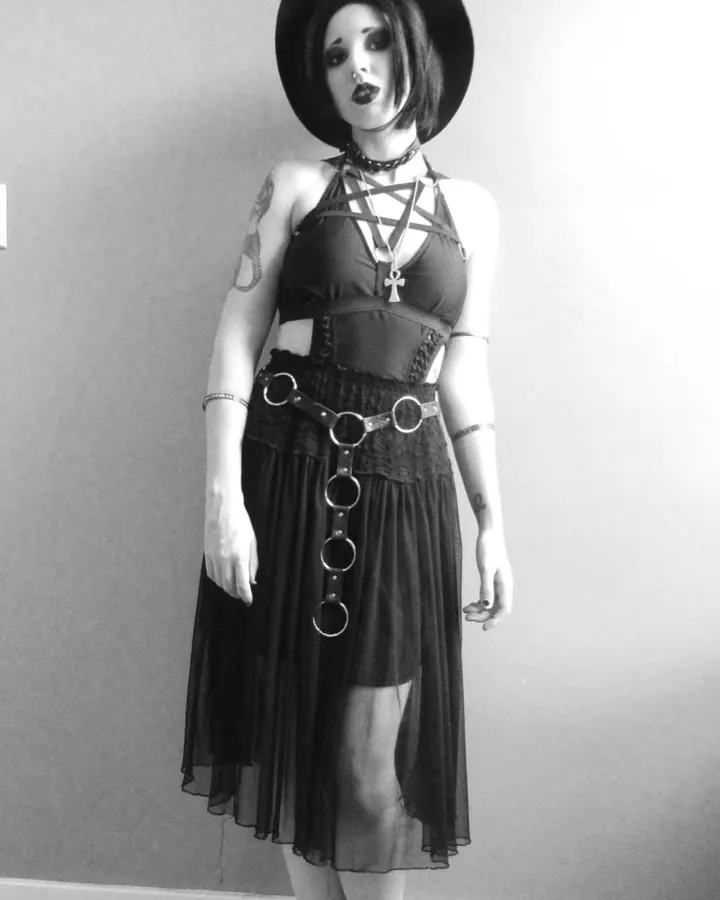Femme blanche aux cheveux noirs, vêtue d'une robe noire avec des anneaux métalliques, chapeau large, regard intense, photo en noir et blanc.