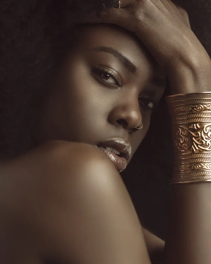 Femme africaine aux cheveux bouclés, regard intense, main sur le front, bracelet doré orné de motifs.