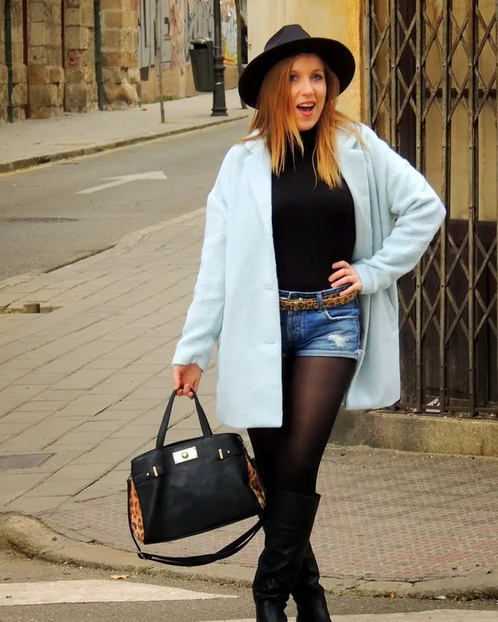 Femme blonde aux cheveux mi-longs, vêtue d'un manteau bleu clair, short en jean, bottes noires, tenant un sac noir, souriant, rue en arrière-plan.