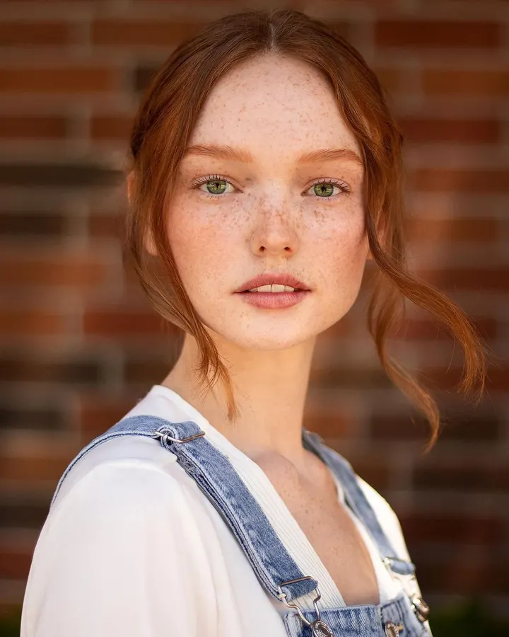 Femme aux cheveux bruns, portant un haut blanc, posant dans une chambre avec un tapis beige et une plante verte.