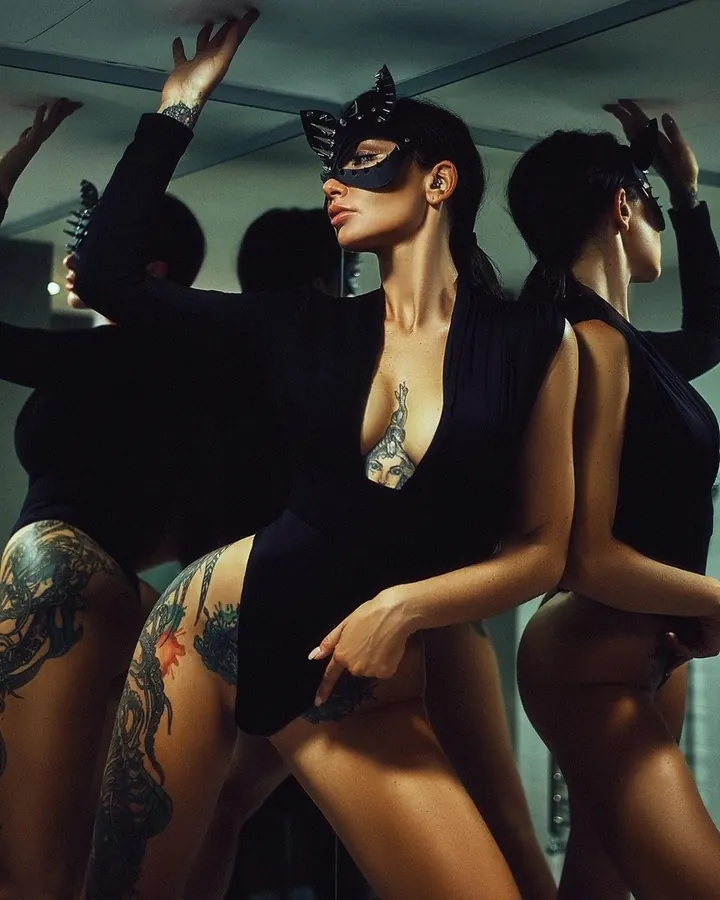 Femme latine en tenue noire, masque de chat, tatouages visibles, posant devant un miroir.