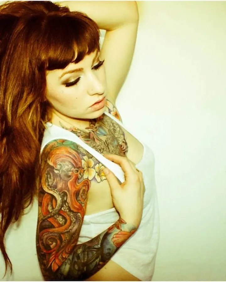 Femme aux cheveux roux, tatouage coloré sur le bras, vêtue d'un haut blanc, main sur la poitrine, regard baissé.
