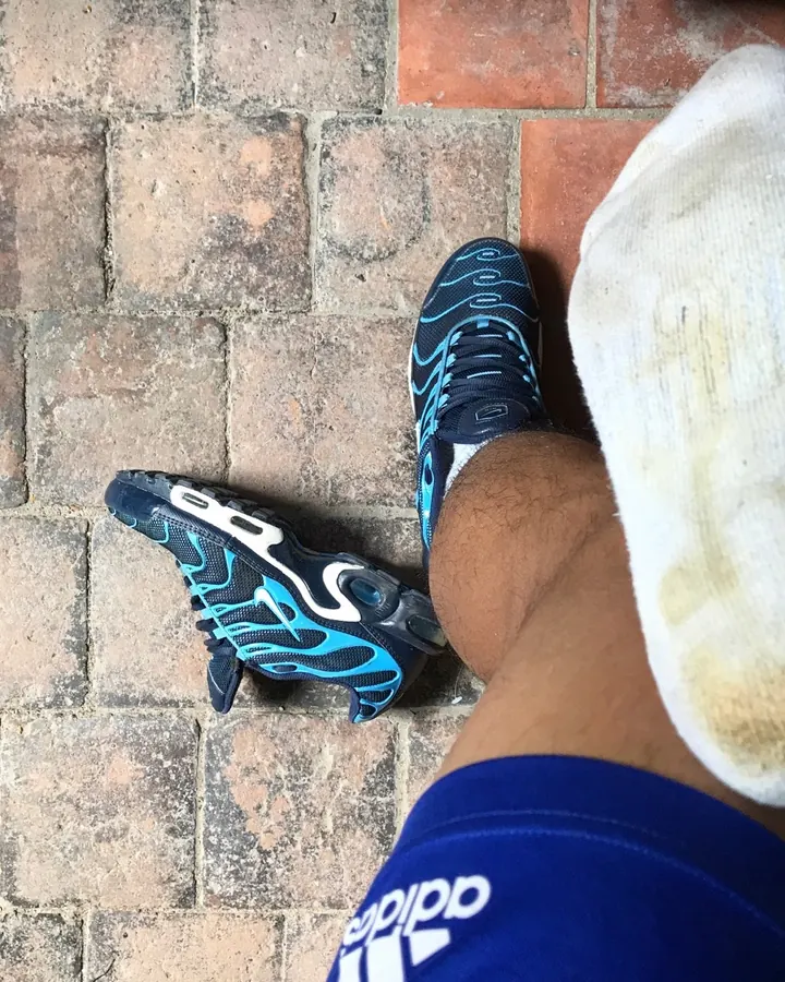 Pieds et chevilles d'un homme portant des chaussures de sport bleues et noires, une chaussette blanche sale, sur un trottoir pavé.