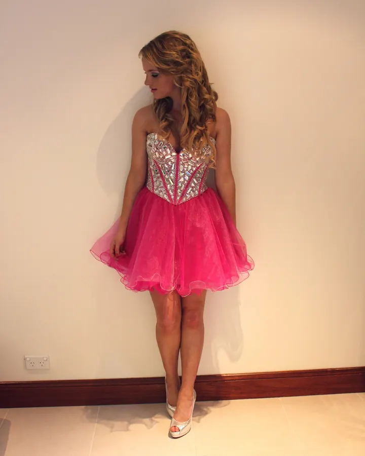 Femme blonde en robe rose pailletée courte, debout contre un mur blanc, regard baissé, pieds en talons argentés.