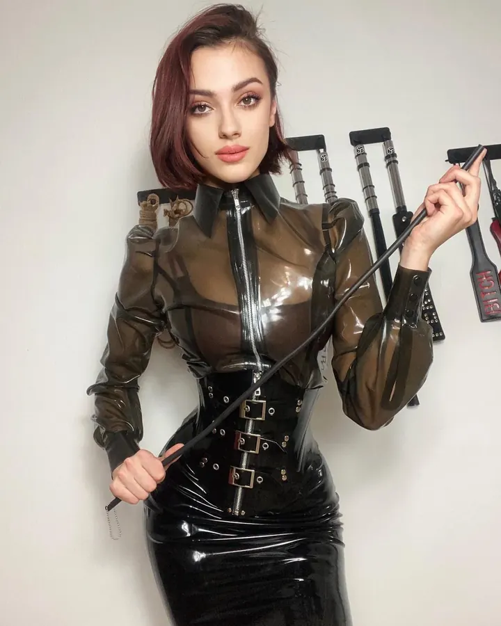 Femme en tenue de latex transparent brun et noir, tenant une cravache, devant un mur blanc avec des accessoires BDSM en arrière-plan.