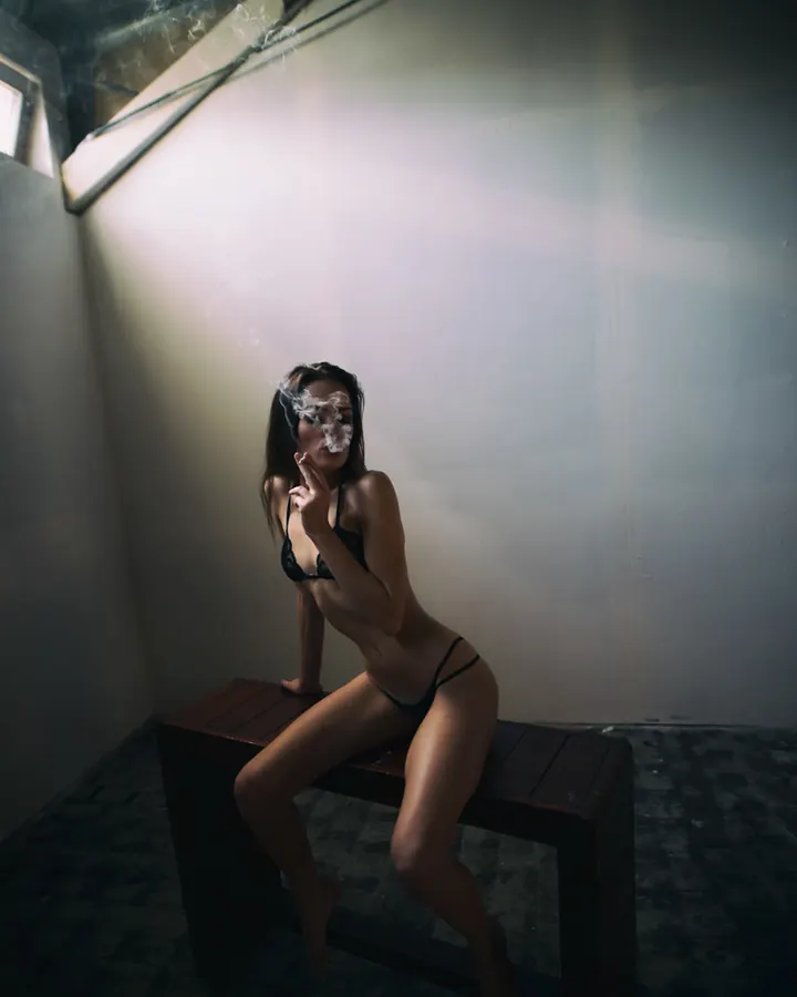 Femme en lingerie noire, assise sur un banc en bois dans une pièce sombre, fumant une cigarette, fumée autour du visage.