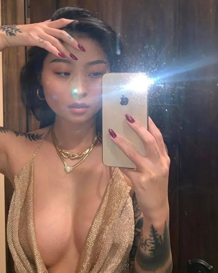 Femme aux cheveux noirs, portant un haut doré et des bijoux, prenant un selfie dans un miroir.