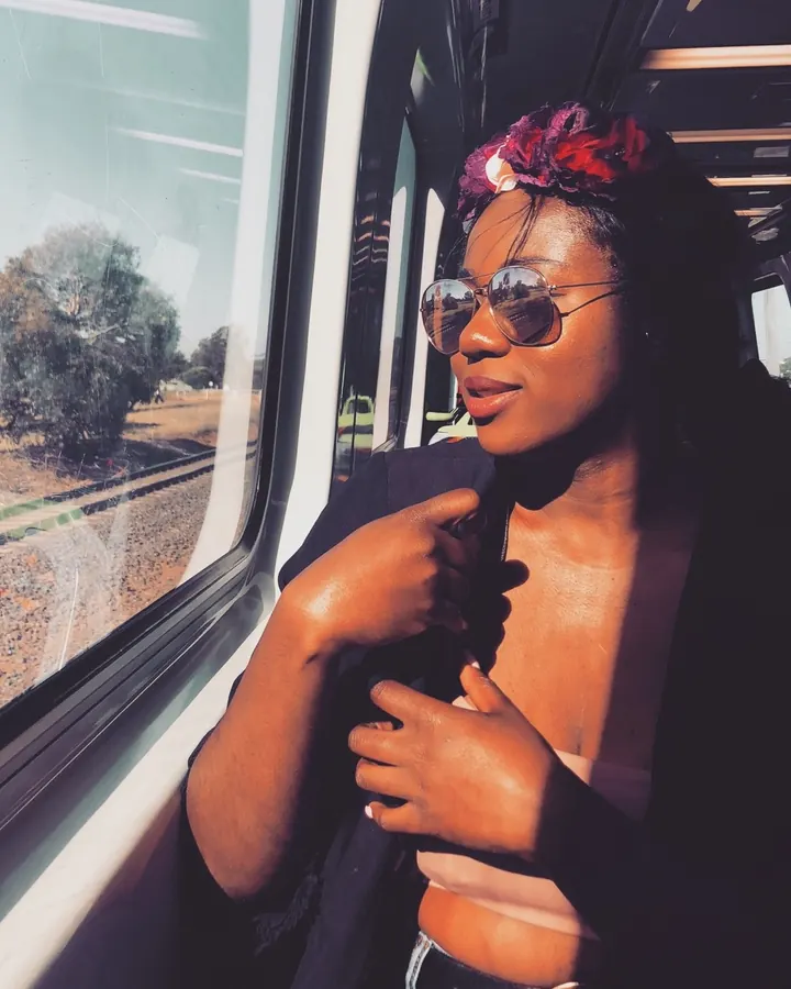 Femme africaine portant des lunettes de soleil et une couronne de fleurs, assise près d'une fenêtre de train, regardant vers l'extérieur.