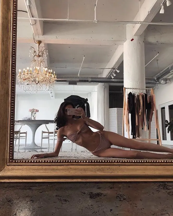 Femme aux cheveux noirs, portant un haut noir et un string noir, posant dans une chambre.