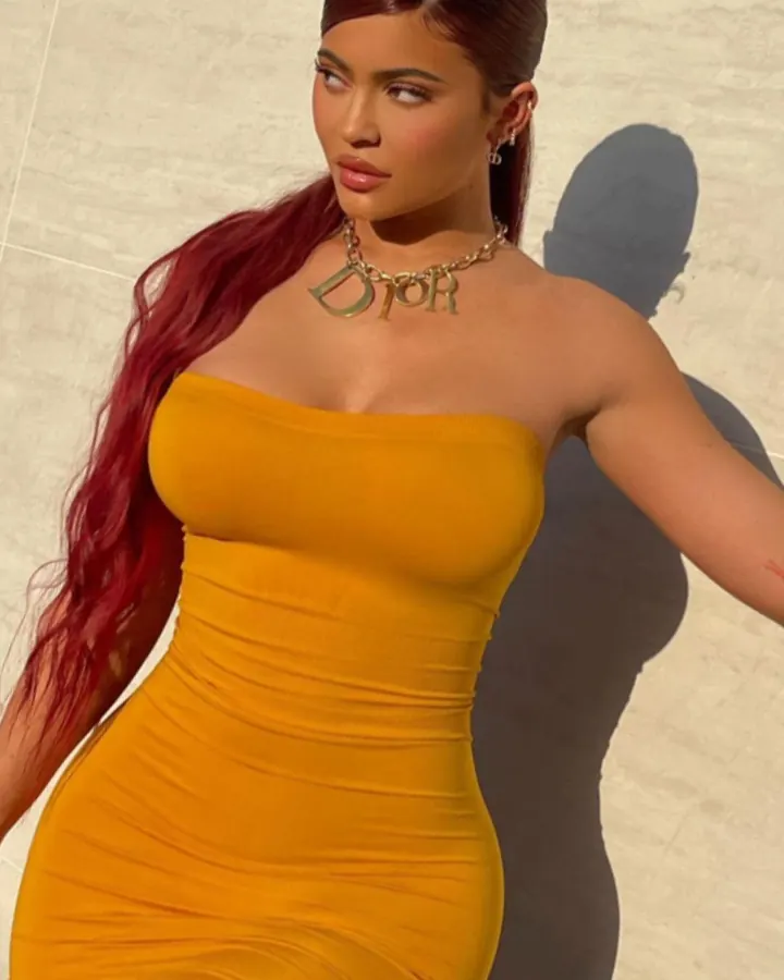 Femme en robe jaune moulante, cheveux rouges, posant contre un mur beige.