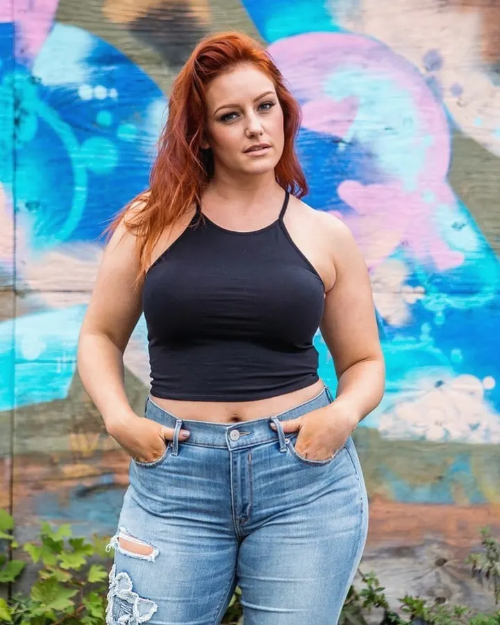 Femme aux cheveux roux, en haut noir et jeans déchirés, devant un mur de graffiti coloré.