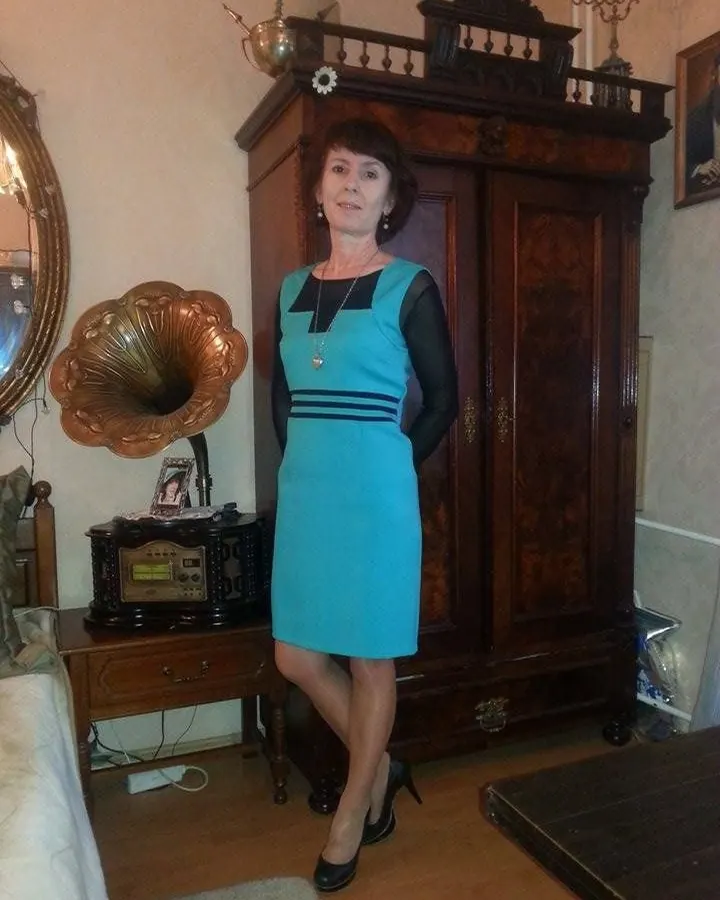 Femme d'âge mûr en robe bleue, debout près d'un meuble ancien avec un gramophone, dans une chambre classique.