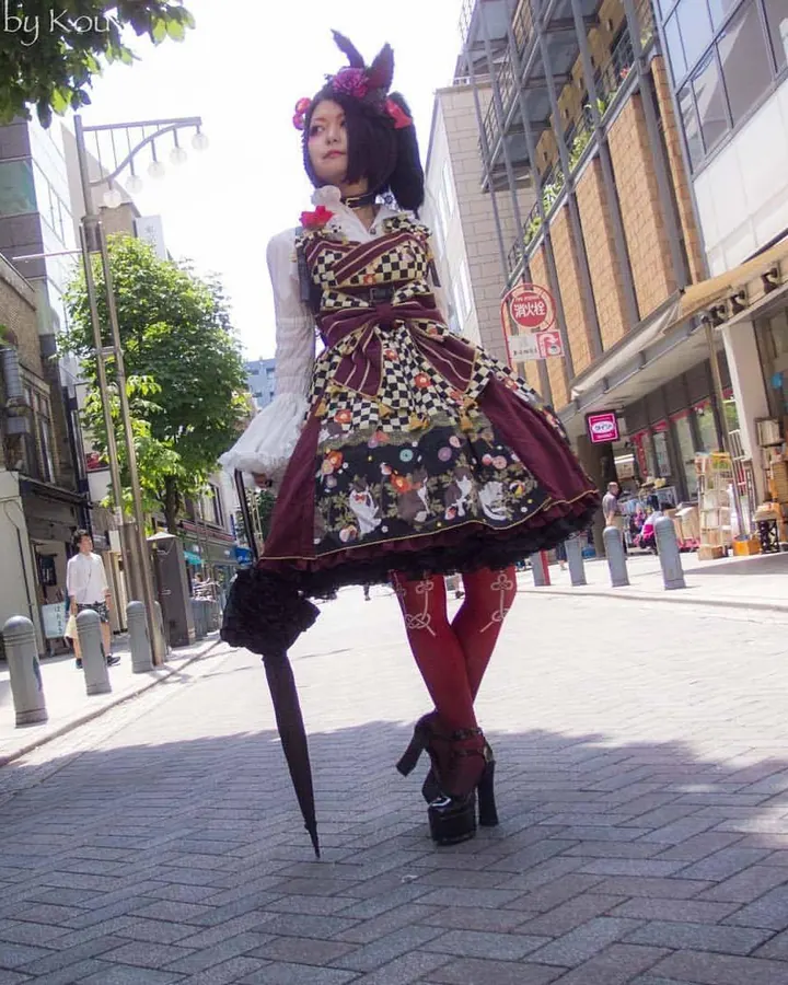 Jeune femme asiatique en robe Lolita colorée avec chapeau, tenant un parapluie, sur un trottoir urbain ensoleillé.