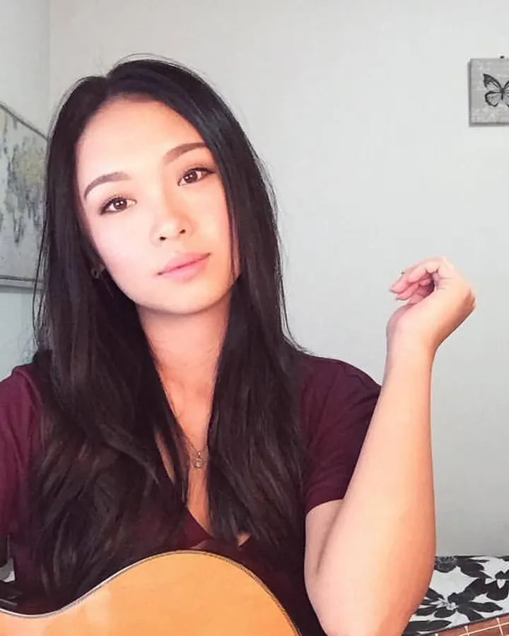 Jeune femme asiatique assise, tenant une guitare, regardant l'appareil, dans une chambre décorée de photos et d'une carte du monde.