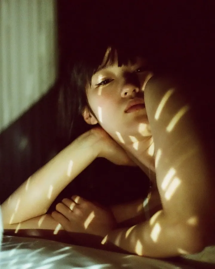 Jeune femme asiatique allongée dans une pièce sombre, lumière projetée sur son visage et son corps, vêtue d'un haut blanc.
