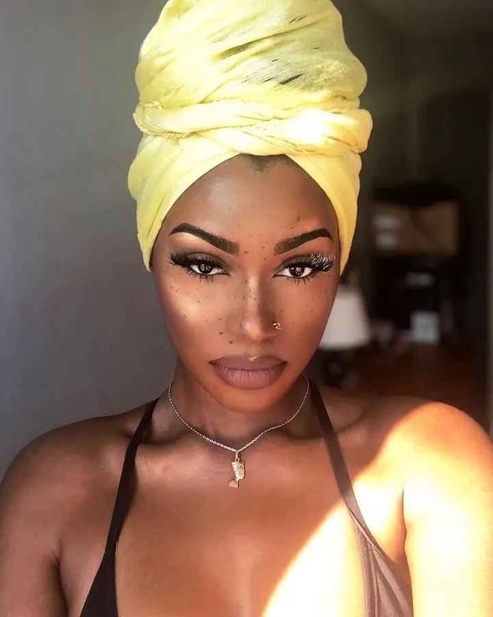 Femme africaine au teint foncé portant un turban jaune, maquillage prononcé, collier fin, regard direct.