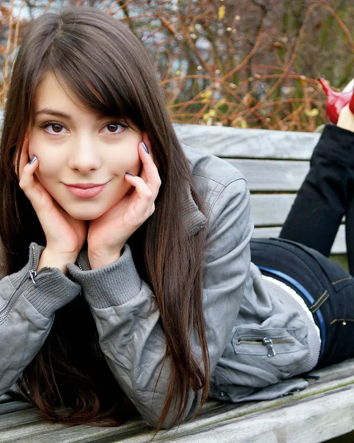 Jeune femme aux cheveux bruns allongée sur un banc en bois, souriante, mains sous le menton, chaussures rouges.