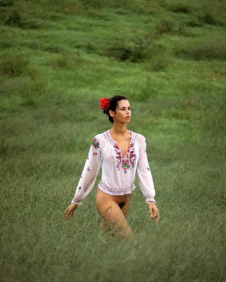 Jeune femme aux cheveux bruns-noirs portant une chemise blanche brodée, marchant dans un champ vert avec une fleur rouge dans les cheveux.