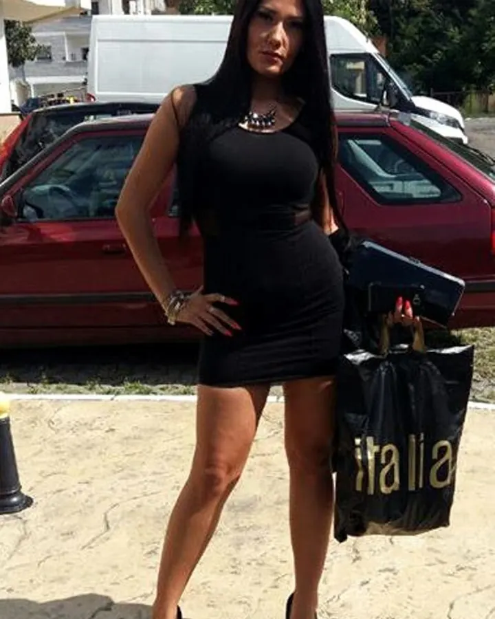 Femme aux cheveux noirs, portant un corset noir en latex, les mains sur les hanches, posant devant un mur beige.