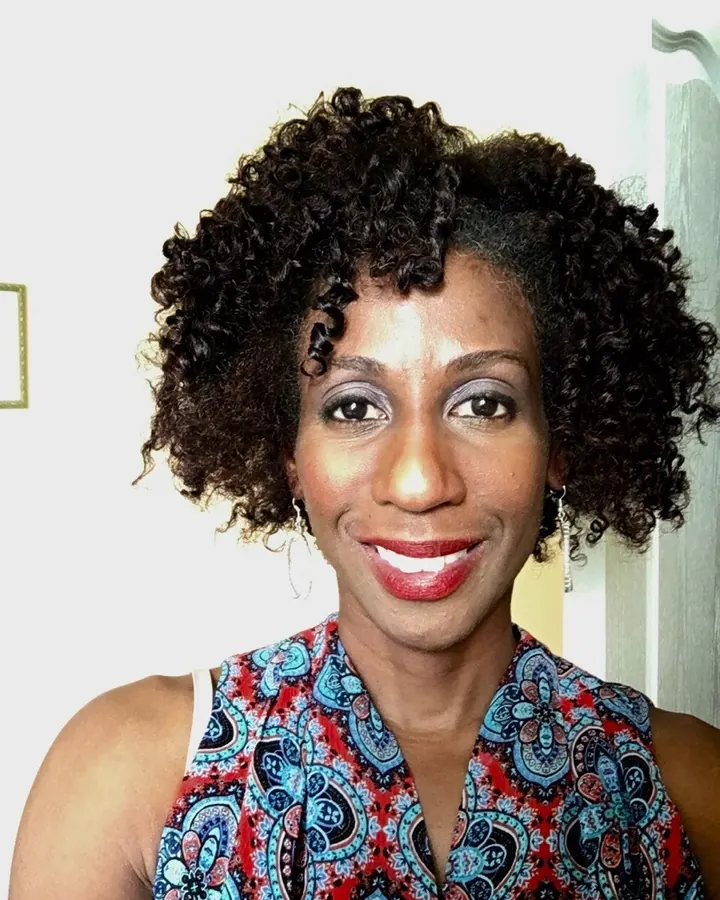 Femme afro aux cheveux bouclés, portant un haut violet et blanc, posant dans une pièce avec une lumière chaude.
