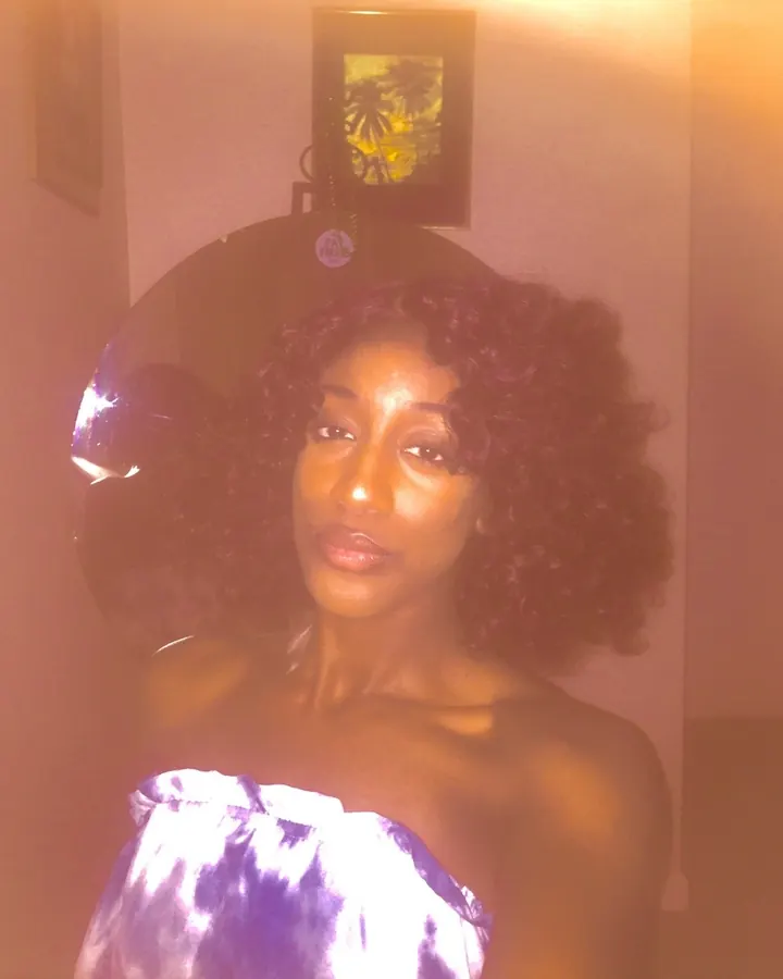 Femme afro aux cheveux bouclés, portant un haut violet et blanc, posant dans une pièce avec une lumière chaude.