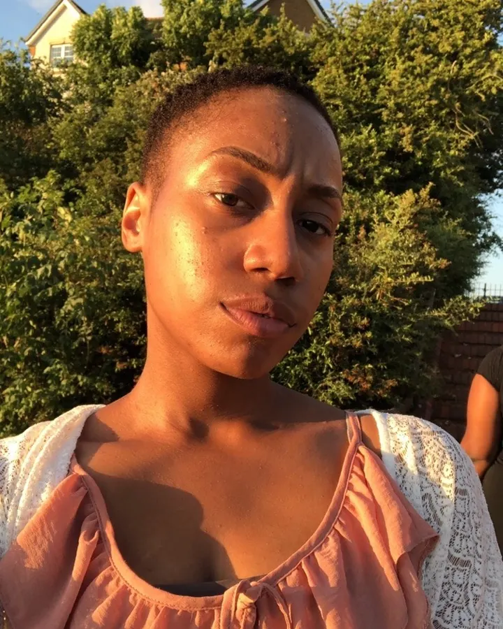 Femme afro aux cheveux courts, portant un haut rose et un cardigan blanc, souriant légèrement sous la lumière du soleil.