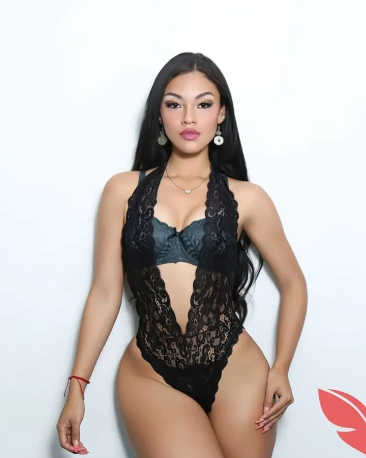 Femme latine aux longs cheveux noirs, vêtue d’un body noir en dentelle, posant devant un fond blanc.