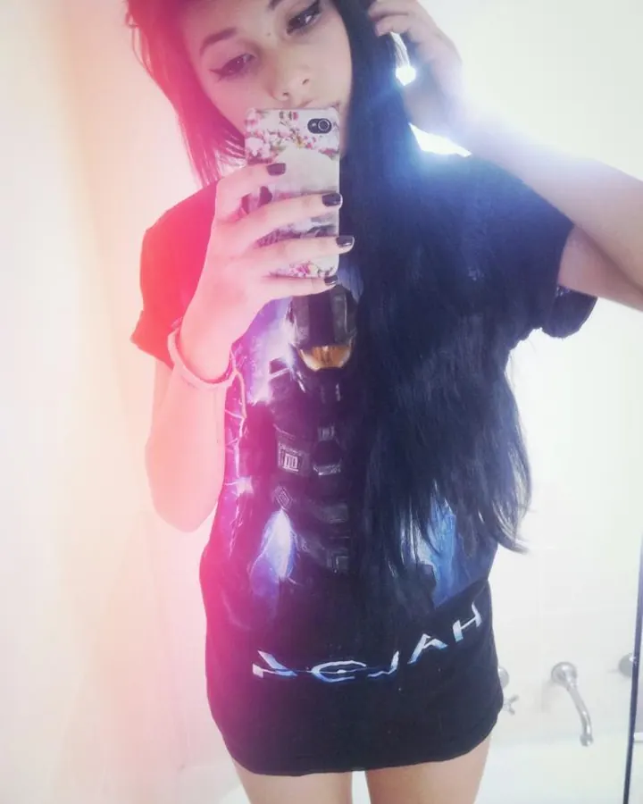 Femme asiatique aux longs cheveux noirs, vêtue d'un t-shirt noir graphique, prenant un selfie dans un miroir.