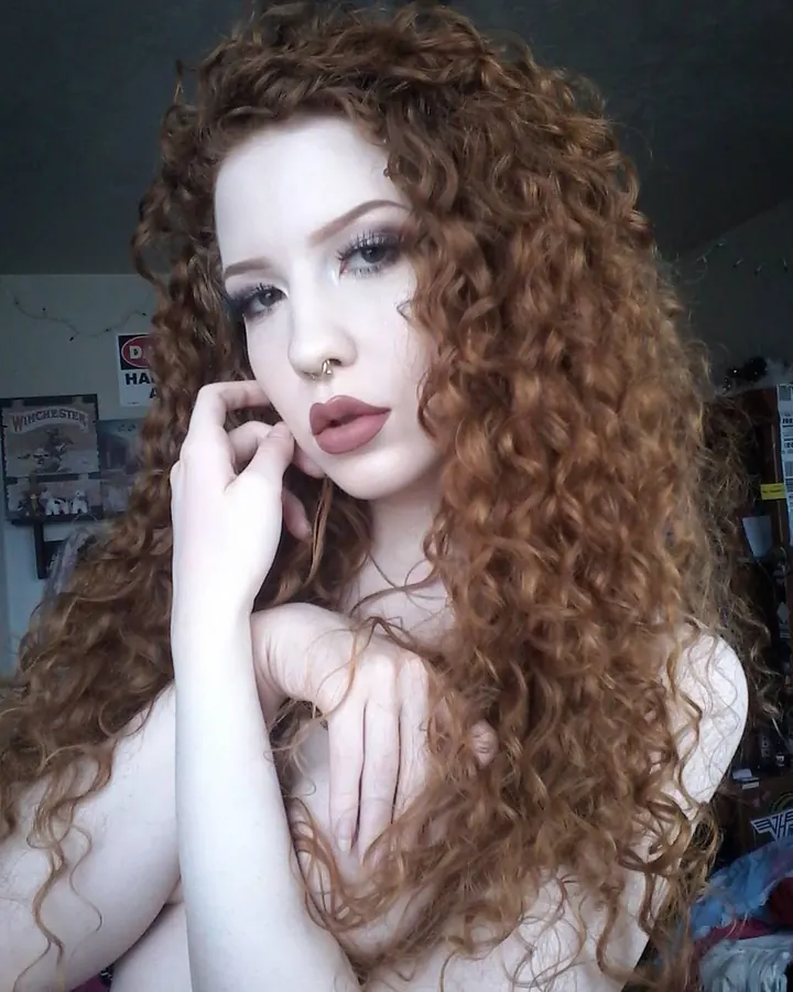 Femme aux cheveux roux bouclés, maquillage prononcé, regard intense, main près du visage.