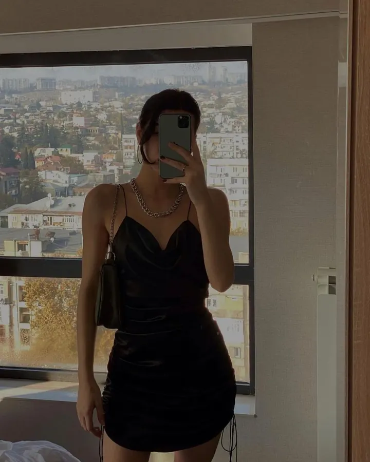 Femme aux cheveux bruns foncés, portant une robe noire en cuir synthétique, prenant un selfie devant une fenêtre avec une vue sur une ville, tenant son téléphone devant son visage.
