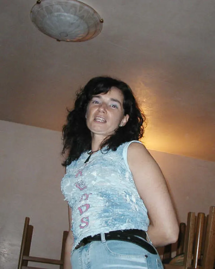 Femme aux cheveux bruns foncés, portant un débardeur bleu clair avec l'inscription 'SPEEDS', une ceinture noire et un jean, souriant largement dans une pièce intérieure.