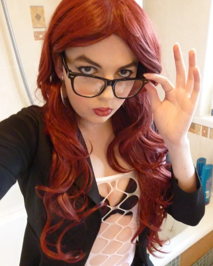 Femme aux cheveux roux longs, portant des lunettes noires, un blazer noir et un haut blanc ajouré, tenant ses lunettes avec la main droite, dans une salle de bains.