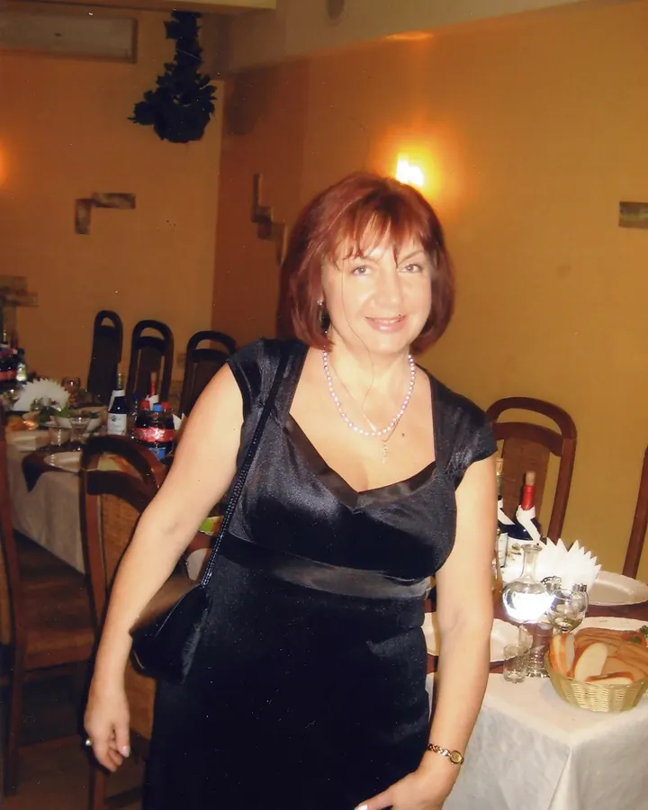 Femme aux cheveux bruns foncés, portant une robe lilas courte, debout dans une chambre, les mains sur les hanches, regardant l'objectif.