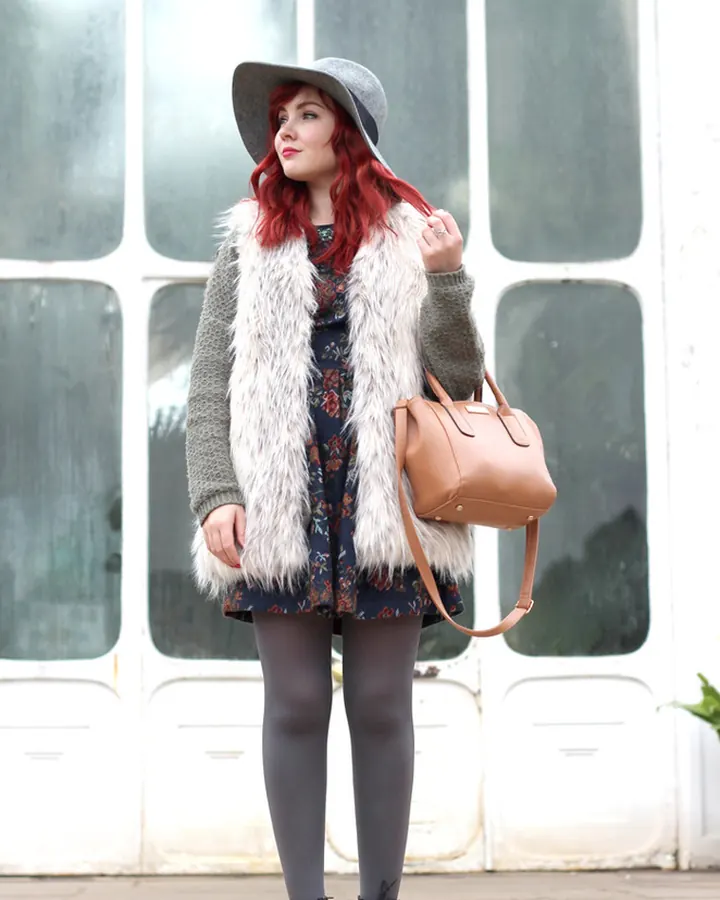 Femme aux cheveux roux portant un chapeau gris, une veste en fausse fourrure blanche, une robe fleurie, des collants gris et des bottines noires, debout devant une porte vitrée
