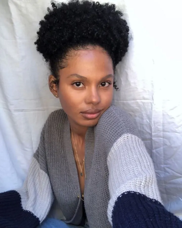 Femme aux cheveux noirs bouclés portant un cardigan tricoté gris et bleu, assise sur un lit blanc