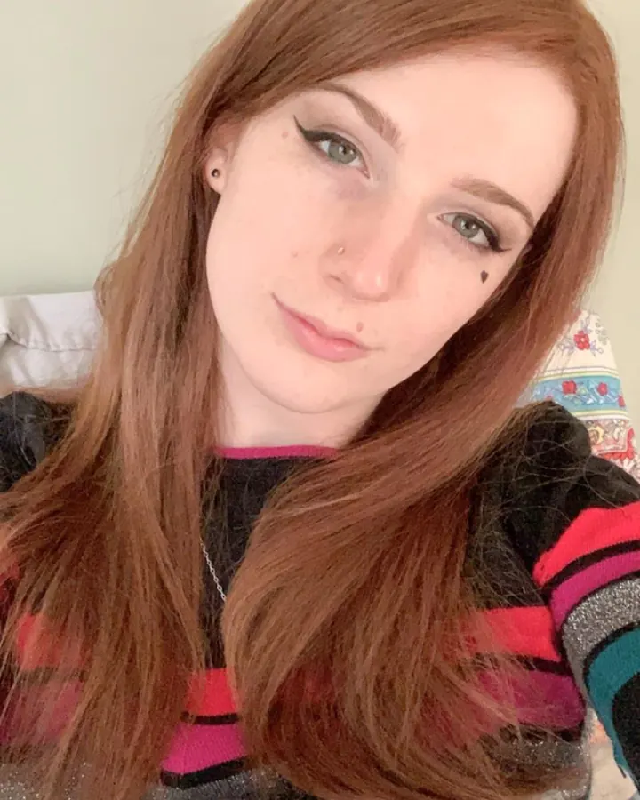 Femme aux cheveux roux portant un haut noir, posant dans une chambre avec un lit en arrière-plan