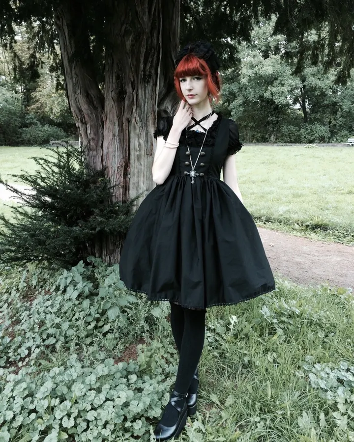 Jeune femme aux cheveux roux, en robe noire gothique, posant devant un arbre dans un parc.