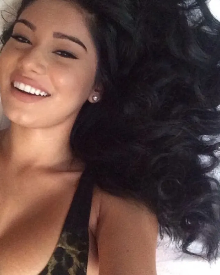 Femme latine aux cheveux bruns-noirs, souriante, allongée sur un lit blanc, en haut léopard.