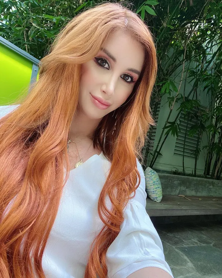 Femme aux cheveux roux longs, maquillage soigné, vêtue d'une chemise blanche, en extérieur devant des bambous.