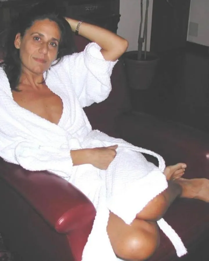 Femme latine aux cheveux bruns-noirs, en haut noir, posant sur un lit blanc.