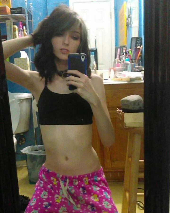 Jeune femme aux cheveux bruns-noirs, en sous-vêtement noir et pantalon rose à motifs, prenant un selfie dans une salle de bain bleue.
