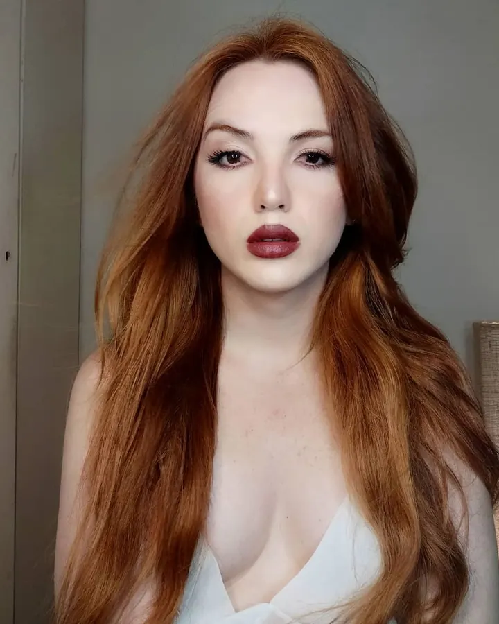 Femme aux cheveux roux longs, portant une robe blanche à col en V, regardant directement l'appareil photo.
