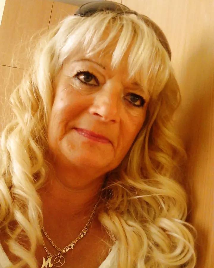 Femme aux cheveux blonds portant un haut blanc et un short blanc, appuyée contre un arbre près d'un étang.