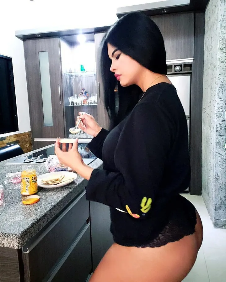 Femme aux cheveux noirs portant un haut rose et un legging noir, posant près d'une table blanche devant une porte en bois.