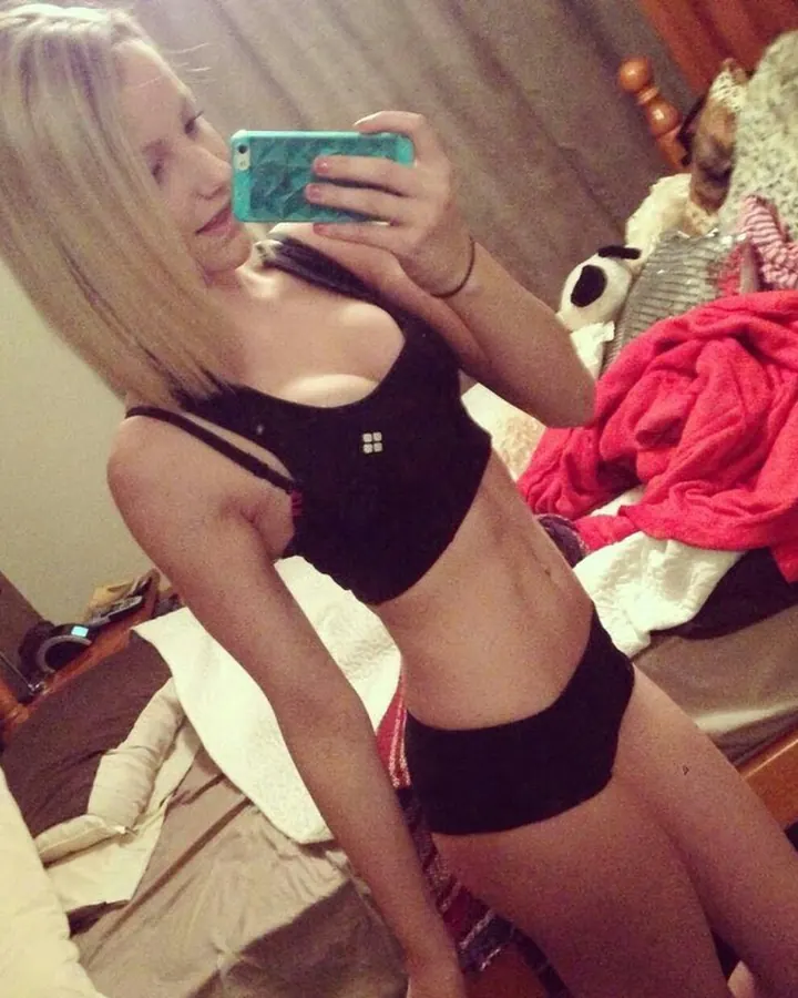 Femme blonde aux cheveux courts portant un haut noir et un string noir, prenant un selfie dans une chambre.