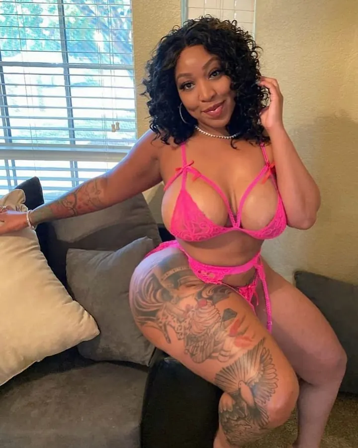Femme aux cheveux noirs en bikini rose, assise sur un canapé, montrant un tatouage sur la cuisse.
