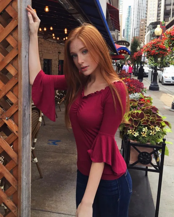 Femme aux cheveux roux en haut rouge, appuyée contre un poteau sur un trottoir animé.